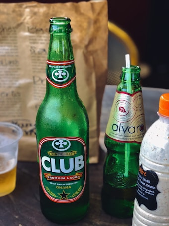 A table display featuring an assortment of beverages. There is a large green glass bottle of Club Premium Lager from Ghana, adorned with branding labels. Next to it is a slightly smaller bottle of Alvaro in a lighter green hue with decorative labeling. Also present is a plastic bottle containing a creamy-looking drink with a label listing ingredients like milk, vanilla, and cinnamon. In the background, a brown paper bag, possibly for takeout, is visible along with a partially visible plastic cup containing a light-colored liquid.
