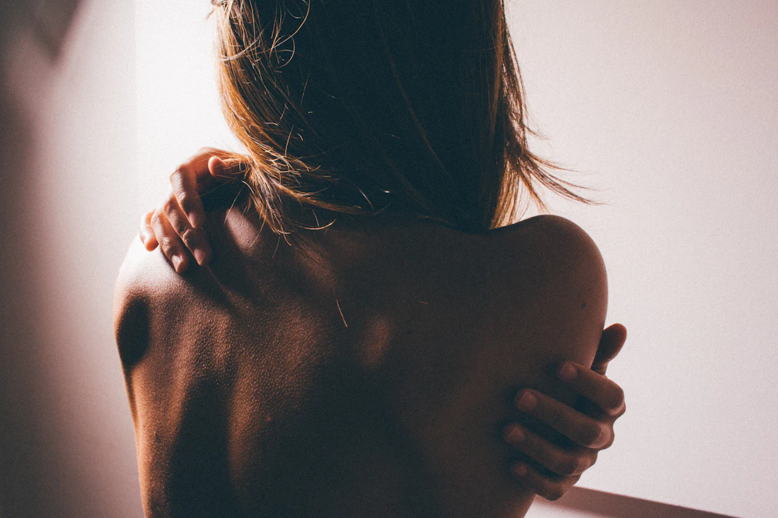 A woman's bare back and shoulder from behind, hair falling over, one arm across her chest; no face