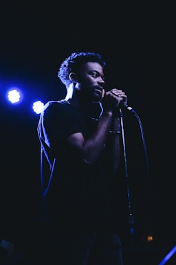 A person is standing in a dimly lit environment, holding a microphone close to their mouth as if they are singing or speaking. The lighting creates a dramatic shadow across their face, emphasizing their focus and concentration.