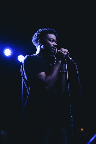 A person is standing in a dimly lit environment, holding a microphone close to their mouth as if they are singing or speaking. The lighting creates a dramatic shadow across their face, emphasizing their focus and concentration.