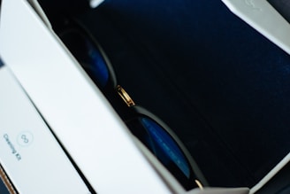 A pair of sunglasses resting inside a box lined with dark blue fabric. The sunglasses feature a metal bridge with black and gold details. The interior of the box is partially visible with a section labeled 'Cleaning Kit' on the inside.