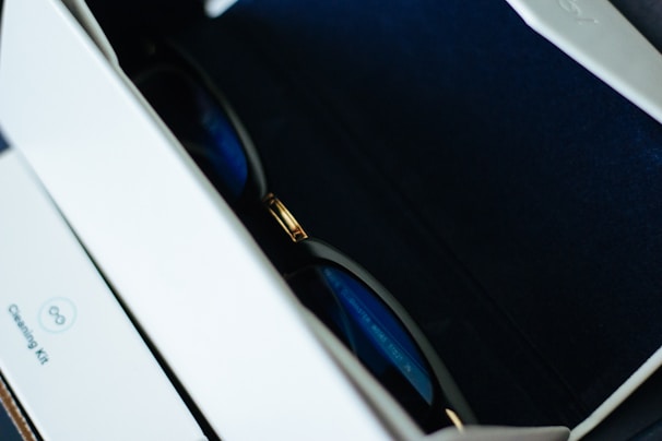 A pair of sunglasses resting inside a box lined with dark blue fabric. The sunglasses feature a metal bridge with black and gold details. The interior of the box is partially visible with a section labeled 'Cleaning Kit' on the inside.