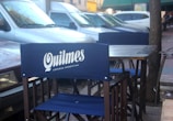 A street-side cafe setup with blue chairs branded with Quilmes, an Argentinian beer. Parked cars are in the background next to a sidewalk and trees.