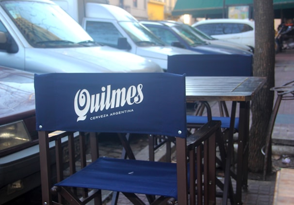 A street-side cafe setup with blue chairs branded with Quilmes, an Argentinian beer. Parked cars are in the background next to a sidewalk and trees.