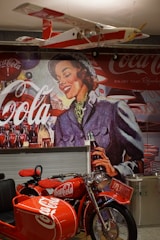A vintage-style mural featuring a smiling woman holding a Coca-Cola bottle, set against a backdrop of Coca-Cola branding. Below the mural, a bright red motorcycle with a sidecar is prominently displayed, also bearing Coca-Cola branding. There is a model airplane hanging from the ceiling, painted in red and white with Coca-Cola logos.