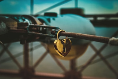 A golden heart-shaped lock is attached to a rusted metal rod. The background appears to be blurred, featuring hints of structural elements, possibly part of a bridge or industrial setting. The lighting gives the image a moody and slightly nostalgic feel.