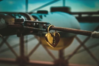 A golden heart-shaped lock is attached to a rusted metal rod. The background appears to be blurred, featuring hints of structural elements, possibly part of a bridge or industrial setting. The lighting gives the image a moody and slightly nostalgic feel.