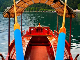 A wooden boat with a canopy over top is positioned on a serene lake. The boat has bright blue paddles and features polished wooden benches and structure. In the background, there are lush green trees lining the shore and a gentle ripple appears on the water's surface.