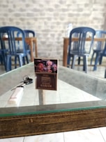 A pack of Gudang Garam cigarettes is placed on a glass-top wooden table. Next to it, there is a white lighter. In the background, several blue plastic chairs are aligned against a wall with a stone-like pattern.