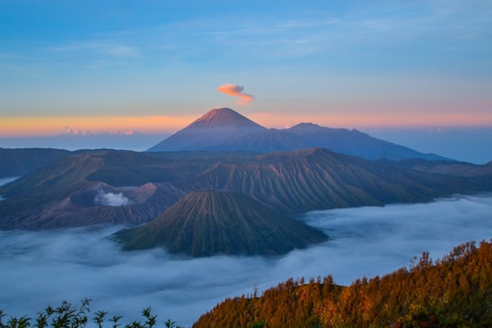 A breathtaking landscape featuring a series of volcanoes under a colorful sky during sunrise or sunset. The scene includes Mount Bromo with a light cloud of smoke and a thin layer of mist or fog that covers the lower areas, creating a mystical atmosphere.