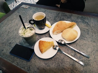 A table with a cup of tea, a green whipped cream-topped beverage in a plastic cup, two plates of baked goods including a slice of cake and two bread rolls. Two metal spoons are placed near the plates and there is a smartphone on the table.