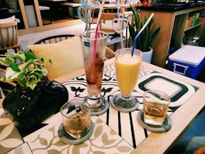A cozy cafe setting with a table featuring two glasses of drinks, a tall iced coffee with pink and blue straws, and a yellow smoothie with a white and blue straw. There are two smaller glasses, each with a translucent liquid, possibly whiskey. A black vase with green leafy plants is placed on the left side of the table. The table has a decorative pattern, and in the background, there are cushioned chairs and indoor plants indicating a relaxed atmosphere.