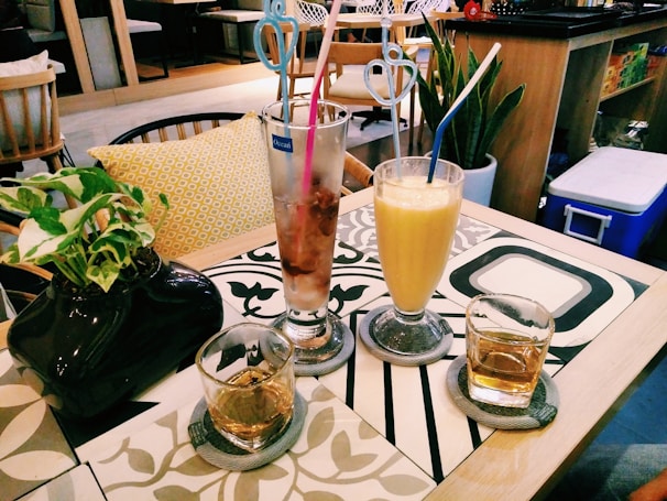 A cozy cafe setting with a table featuring two glasses of drinks, a tall iced coffee with pink and blue straws, and a yellow smoothie with a white and blue straw. There are two smaller glasses, each with a translucent liquid, possibly whiskey. A black vase with green leafy plants is placed on the left side of the table. The table has a decorative pattern, and in the background, there are cushioned chairs and indoor plants indicating a relaxed atmosphere.