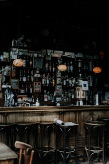 A dimly lit bar interior with a wooden counter and high stools. Various liquor bottles are displayed on shelves behind the bar, accompanied by an assortment of posters and signs. Warm, glowing lights hang above, casting a cozy ambiance.
