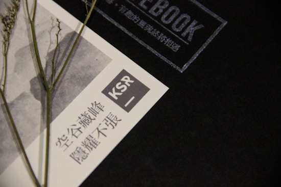 A black notebook cover with the word 'NOTEBOOK' and additional text in Chinese. On the left side, there is a printed piece of paper featuring Chinese characters and the acronym 'KSR'. A dried plant is laid across the paper and notebook.