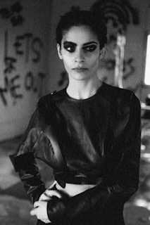 A person with dramatic, bold makeup and an intense expression, dressed in a dark, avant-garde outfit. The setting appears to be urban and edgy, with graffiti on the walls in the background.