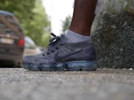 A close-up view of an athletic shoe worn by a person standing on a textured pavement. The shoe features a dark grey knit design with transparent cushioning in the sole. A blurred background shows part of a car and green foliage.
