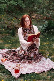 A woman with red hair and glasses is sitting on a patterned blanket in a grassy area, reading a book with a red cover. She is wearing a floral dress with white sleeves. Beside her on the blanket are a pitcher filled with red liquid, a partially eaten watermelon slice, and small colorful jars.