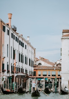 A picturesque canal scene with gondolas navigating the waterway alongside classic Venetian architecture. The buildings feature a mix of white and beige facades with dark shutters and terracotta roofs. Flower boxes adorn some windows, and a gondolier is seen steering a gondola, adding a traditional touch to the serene waterway.