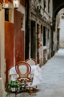 A narrow cobblestone alleyway features a small dining table with a white tablecloth and two wooden chairs. The alley is lined with weathered buildings in earthy tones and a hanging lantern provides a warm glow.