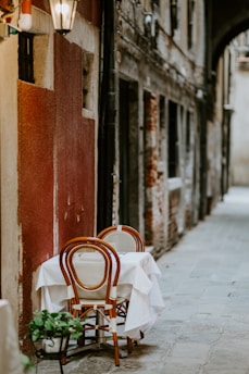 A narrow cobblestone alleyway features a small dining table with a white tablecloth and two wooden chairs. The alley is lined with weathered buildings in earthy tones and a hanging lantern provides a warm glow.
