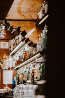A cozy bar interior with shelves full of various liquor bottles. The warm wooden decor and soft lighting create a welcoming atmosphere. Glasses are neatly stacked at the counter, and the arrangement highlights the diversity of beverages available.