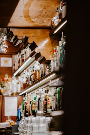 A cozy bar interior with shelves full of various liquor bottles. The warm wooden decor and soft lighting create a welcoming atmosphere. Glasses are neatly stacked at the counter, and the arrangement highlights the diversity of beverages available.