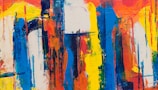 An abstract painting with bold and energetic brushstrokes, featuring a mix of vibrant colors including blue, orange, yellow, and white. The arrangement is chaotic yet harmonious, with drips and smudges that create a sense of movement and depth.