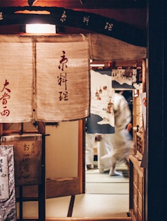 A traditional Japanese entrance with fabric drapes featuring Kanji characters. People in traditional attire are seen through the doorway, creating a sense of movement. The area is dimly lit, and the interior includes wooden elements with tatami mats on the floor.