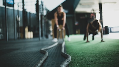 Two individuals are engaging in an intense workout session on a green artificial turf in a gym setting. One person appears to be holding and using battle ropes, while another is performing a plank or push-up exercise nearby. The gym environment includes exercise equipment and a spacious, industrial-style interior.