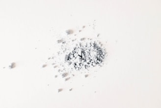 A small pile of gray powder and crushed fragments is scattered on a light surface. The particles are irregular in shape and vary in size, with some larger pieces near the center.