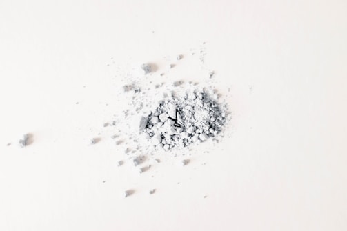 A small pile of gray powder and crushed fragments is scattered on a light surface. The particles are irregular in shape and vary in size, with some larger pieces near the center.