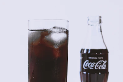A close-up view of a glass filled with a dark, fizzy liquid and ice cubes. Next to the glass, there is a Coca-Cola bottle with the original taste label visible on it.