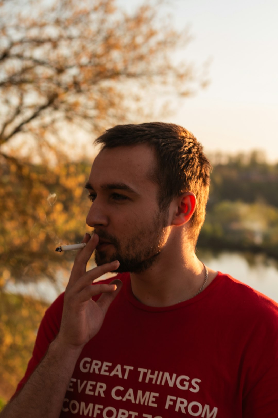 man standing smoking cigarette near body of water and trees