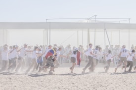 A group of people participate in an outdoor activity on a sandy surface, with many wearing matching T-shirts and hats. Some are draped in flags, and they appear to be moving in a chain-like formation. The background features an overhead structure with seated spectators.