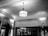 A black and white image of an elegant interior space with a classic ceiling and a central vintage-style hanging lamp. Decorative molding lines the ceilings and walls, complemented by curtains and tall ornamental plants. The lighting casts intricate shadows on the ceiling.