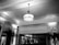 A black and white image of an elegant interior space with a classic ceiling and a central vintage-style hanging lamp. Decorative molding lines the ceilings and walls, complemented by curtains and tall ornamental plants. The lighting casts intricate shadows on the ceiling.