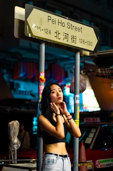 A woman is standing under a street sign for Pei Ho Street at night. She is wearing a black crop top and light denim jeans. The background is filled with colorful, blurred lights, and there is a red taxi nearby. The scene appears to be in an urban area with various signs and posters on the signpost.