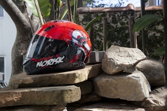 A colorful motorcycle helmet with a red and black design featuring a graphic illustration rests on a pile of large stones. The background shows greenery and a section of a building, suggesting an outdoor setting.