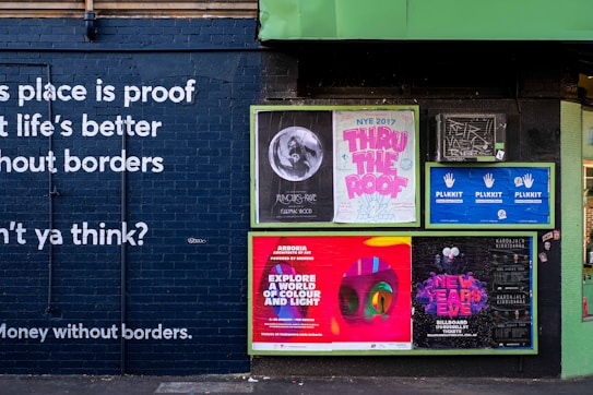 A brick wall is adorned with several vibrant posters promoting events and concepts. Large, bold white text is painted directly onto the dark brick stating a message about life without borders. The posters include colorful and eye-catching graphics, with themes around music events like New Year's Eve and artistic exhibitions.