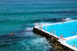 An outdoor swimming pool is seen adjacent to the ocean, where several people are swimming and lounging around. The pool's turquoise water contrasts with the deeper blue of the surrounding sea. Some swimmers are in the ocean while others enjoy the pool, separated by a narrow edge.