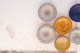Intricately designed ceramic plates are arranged against a textured white wall. The plates feature a variety of colorful and detailed patterns, with central floral or geometric designs. The dominant colors include blues, yellows, oranges, and whites. The arrangement and patterns evoke a traditional or handcrafted aesthetic.
