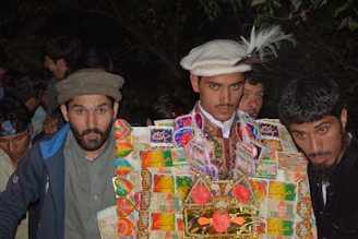 A group of people gathered closely together. The central figure is dressed in a colorful traditional outfit adorned with currency notes and decorative elements, possibly for a festive or cultural celebration. The background shows several other people, mostly young men, seemingly part of the same gathering.