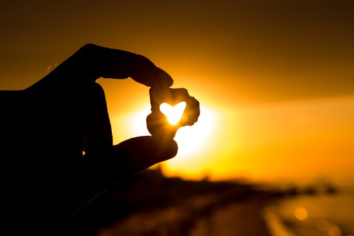 A silhouette of a hand holding a small heart-shaped object against the backdrop of a glowing sunset. The warm colors of the setting sun and sky create a serene and romantic ambiance.