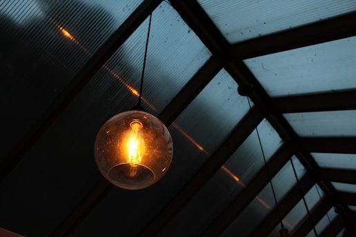 A glowing Edison-style bulb hangs from a ceiling, enclosed in a glass sphere. The background features a grid-like pattern of translucent, blue-tinted panels.