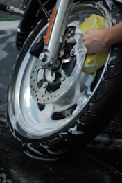 A close-up view of a motorcycle wheel being meticulously cleaned with a sponge. The wheel is covered in soapy water, indicating a thorough wash. The hand holding the sponge appears to be scrubbing the metal rim.