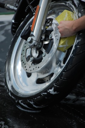 A close-up view of a motorcycle wheel being meticulously cleaned with a sponge. The wheel is covered in soapy water, indicating a thorough wash. The hand holding the sponge appears to be scrubbing the metal rim.