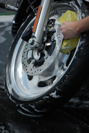 A close-up view of a motorcycle wheel being meticulously cleaned with a sponge. The wheel is covered in soapy water, indicating a thorough wash. The hand holding the sponge appears to be scrubbing the metal rim.