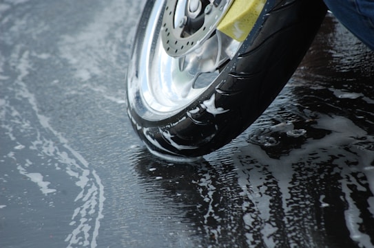 A motorcycle tire with visible soap suds on the wet pavement, suggesting it has been washed. The shiny surface of the wheel adds a reflective quality, contrasting with the dark rubber and the slick, soapy ground beneath.