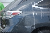 The rear side of a dark-colored vehicle is being washed, as evidenced by streams of white soap and water flowing down the car's surface. Reflections indicate a glossy finish, and part of a person wearing a green shirt is visible in the background.